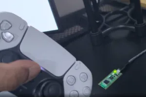 Close-up of a gray PlayStation-style controller connected to a small green circuit board with a glowing green LED on a desk.