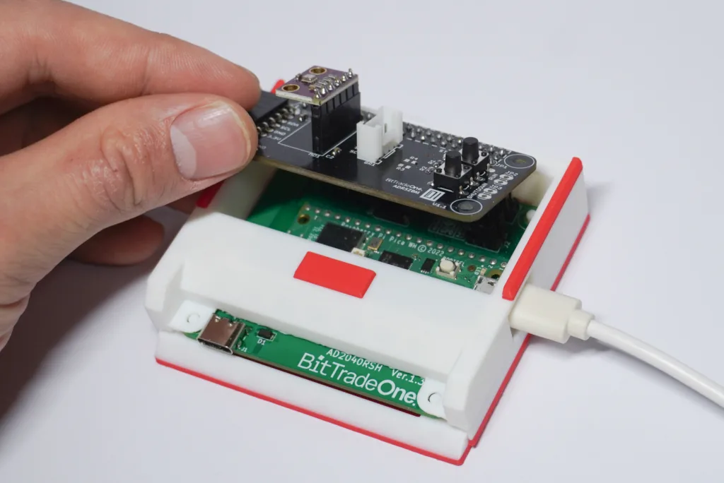 Hand inserting a small PCB module onto a BitTradeOne microcontroller trainer board; USB port visible nearby.