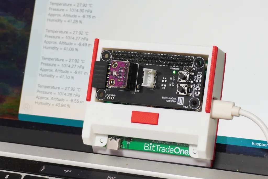 Close-up of a BitTradeOne development board mounted in a white case with red endcaps; a green status LED is lit and multiple connectors are visible.