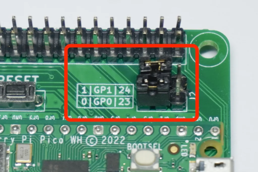 Close-up of a green PCB showing a long row of header pins at the top and a small black jumper switch in the center, with silkscreen labels GP1/GP0 and BOOTSEL nearby.