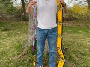 Man outdoors stands by a tree, holding a string of colorful plastic segment pieces and a yellow folding frame.