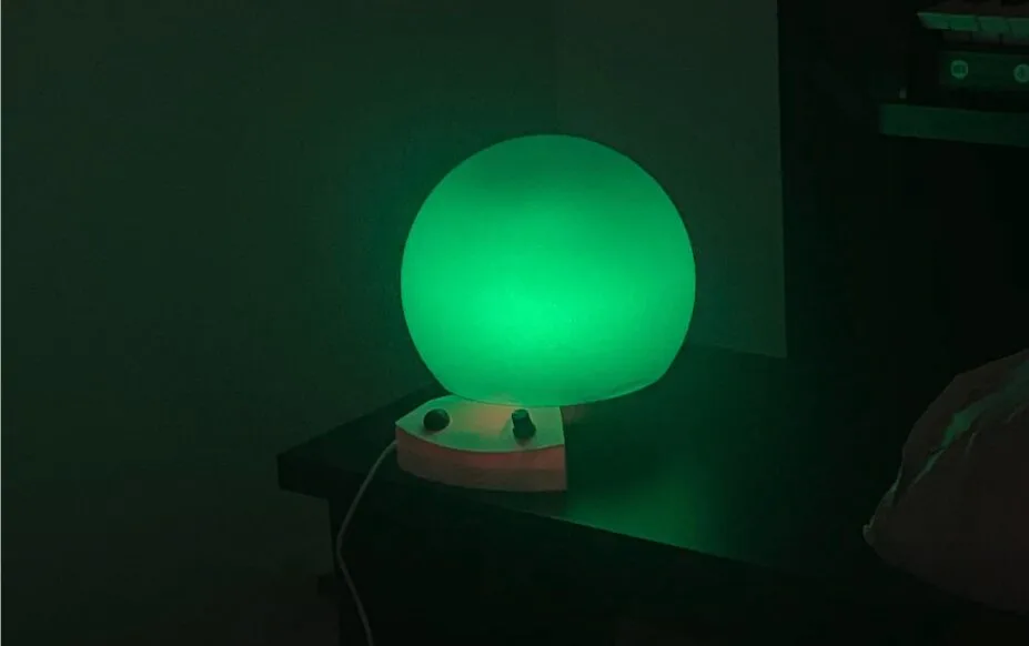 Green glowing orb lamp on a small wooden base sits on a desk in a dark room, casting green light around.
