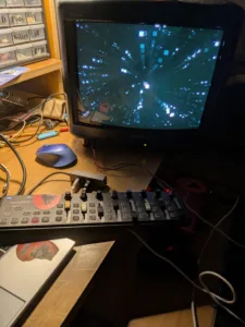 Older CRT monitor displaying a blue digital starburst pattern, with a keyboard and cluttered desk below in a workshop setting.