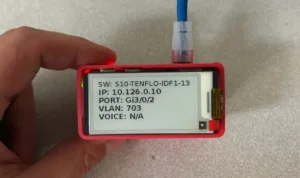 Red handheld network device with a screen showing IP 10.126.0.10, VLAN 703, port Gig0/2 and a blue cable plugged in.
