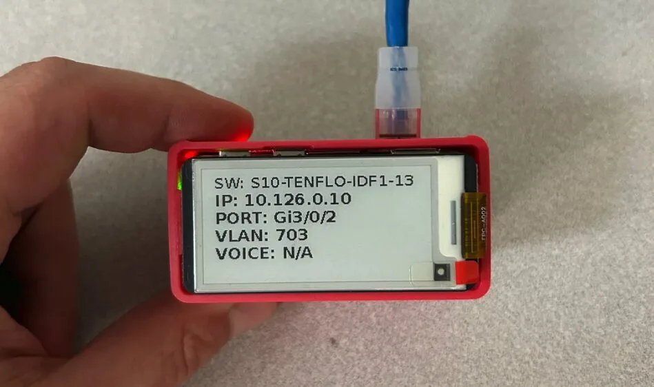 Red handheld network device with a screen showing IP 10.126.0.10, VLAN 703, port Gig0/2 and a blue cable plugged in.