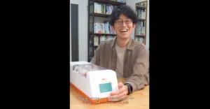 Smiling man with glasses demonstrates a white and orange device on a table, bookshelves in the background.