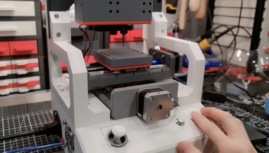 Benchtop CNC machine with a spindle above a metal worktable; a hand rests on the front control panel in a lab/workshop setting.