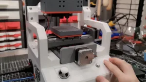 Benchtop CNC machine with a spindle above a metal worktable; a hand rests on the front control panel in a lab/workshop setting.