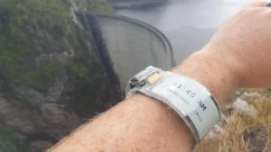Close-up of a damaged silver wristwatch on a hairy forearm, with a rocky, green landscape and a curved dam-like structure in the background.