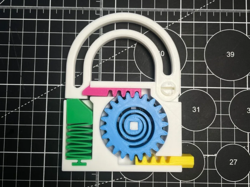 Plastic gear puzzle on a grid-cutting mat: a white base with a large blue gear meshing with inner rings, a green spring at the left, a pink slider above, and a yellow tab at the bottom.
