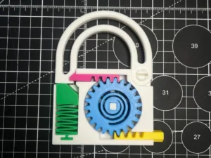 Plastic gear puzzle on a grid-cutting mat: a white base with a large blue gear meshing with inner rings, a green spring at the left, a pink slider above, and a yellow tab at the bottom.