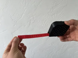 Hands hold a red retractable measuring tape extended, with numbers visible against a white textured wall.