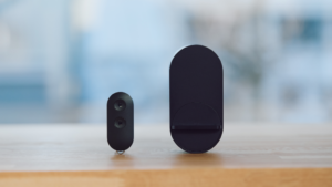 Two small black wireless devices on a wooden table with a soft blue blurred background; a compact oval with two buttons beside a taller rounded base station (likely home security equipment).