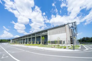 Large industrial building with steel framework and open bays, set beside a curved road under a bright blue sky with clouds; distant snow-capped mountain on the left.