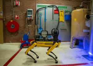 Yellow Boston Dynamics four‑legged robot standing in an industrial workshop with hoses, tools, and safety equipment in view.