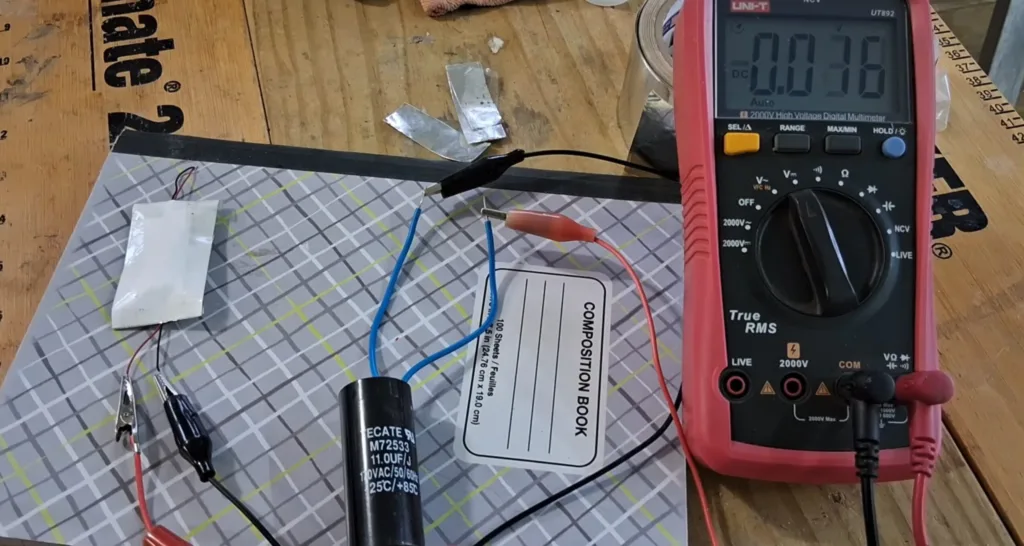 Pink digital multimeter with probes connected to a circuit on a wooden workbench, surrounded by wires and components (capacitor, clips). Rea­ding visible on display is 0.58.