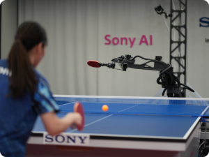 Table tennis table with a robotic arm delivering a shot toward the ball; blurred player on left and 'Sony AI' banner in background.