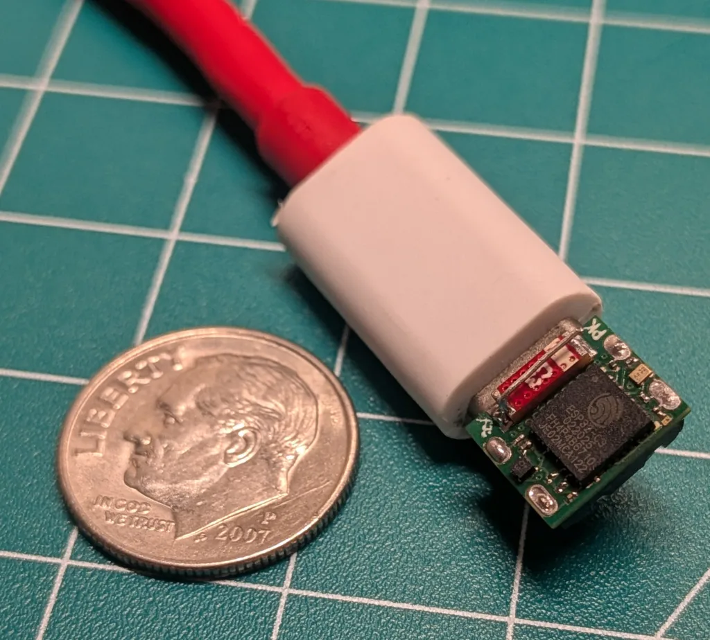 Close-up of a small green circuit board with a red wire attached, partly covered by white heat shrink, next to a U.S. quarter for scale.