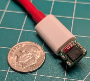 Close-up of a small green circuit board with a red wire attached, partly covered by white heat shrink, next to a U.S. quarter for scale.
