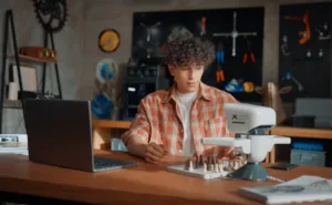 Young man with curly hair in a plaid shirt sits at a workshop desk, writing, with a chessboard and a sewing machine nearby.