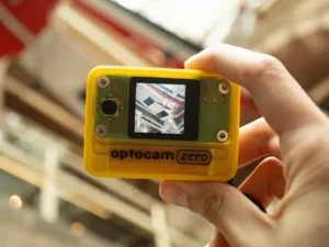 Yellow action camera (Optocam Zero) held in hand, small screen showing a scene being recorded at close range.