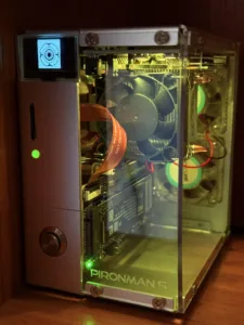 Compact PC with a transparent side panel revealing a cooling fan, orange ribbon cable, PCIe card, and green status LEDs; case labeled PIRANMAN 5 with a small test-pattern display at the top-left edge of the front panel.