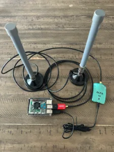 Two tall gray antennas mounted on bases with tangled black cables, connected to a small circuit board; a turquoise Alfa USB wireless adapter nearby.