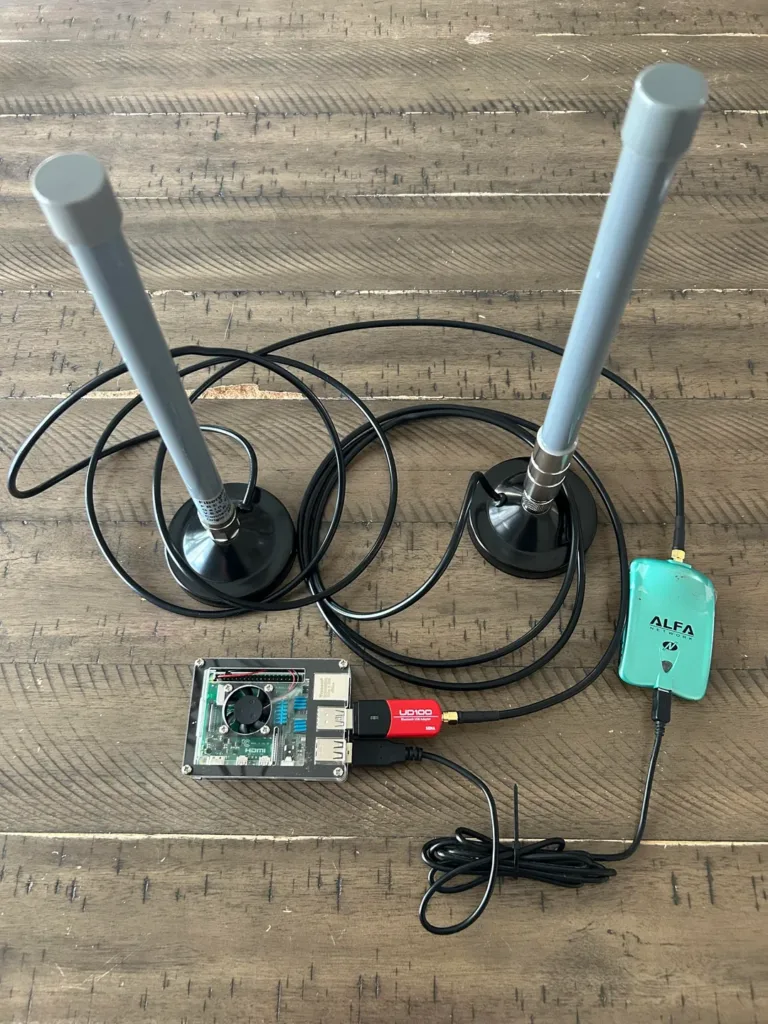 Two tall gray antennas mounted on bases with tangled black cables, connected to a small circuit board; a turquoise Alfa USB wireless adapter nearby.