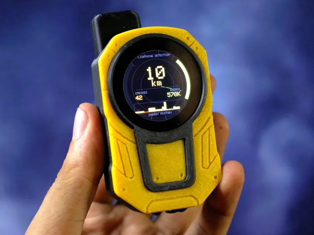 Handheld yellow lightning detector with a round display showing 10 km, 42 strikes, and energy metrics in view.
