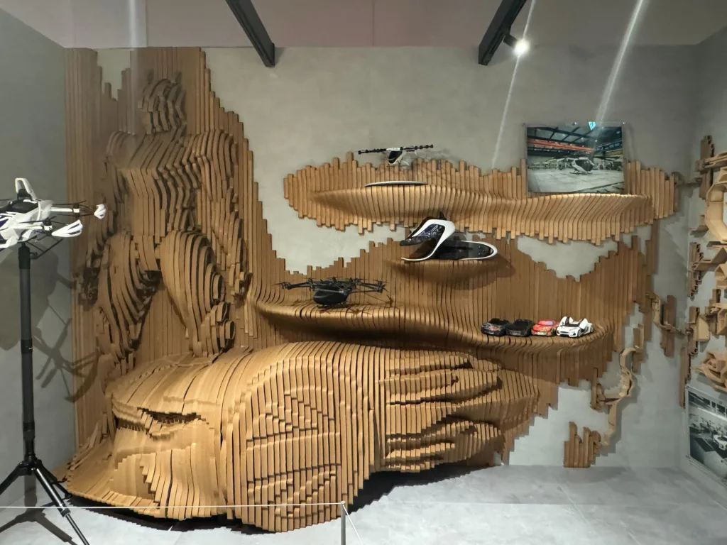 Art installation in a showroom: a wall covered with vertically stacked wooden slats forming a large, wavy wave pattern; several white scooters are displayed on shelves below.