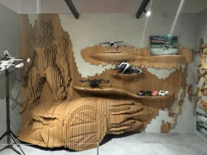 Art installation in a showroom: a wall covered with vertically stacked wooden slats forming a large, wavy wave pattern; several white scooters are displayed on shelves below.