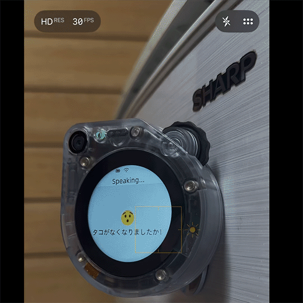 Close-up of a transparent circular translator device mounted on a SHARP panel, screen showing 'Speaking...' and a Japanese message.