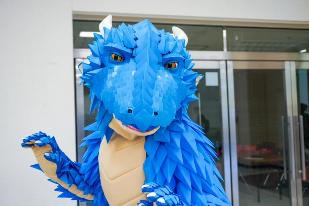 Person in a blue dragon costume with spiky scales standing outdoors near glass doors in a building.