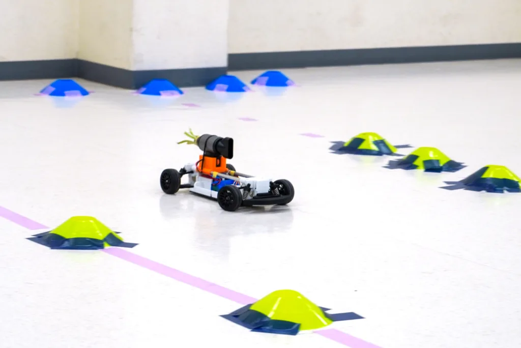 Small programmable robot car driving on a gym floor, passing blue cone markers and neon yellow-green pyramid obstacles in an indoor course.