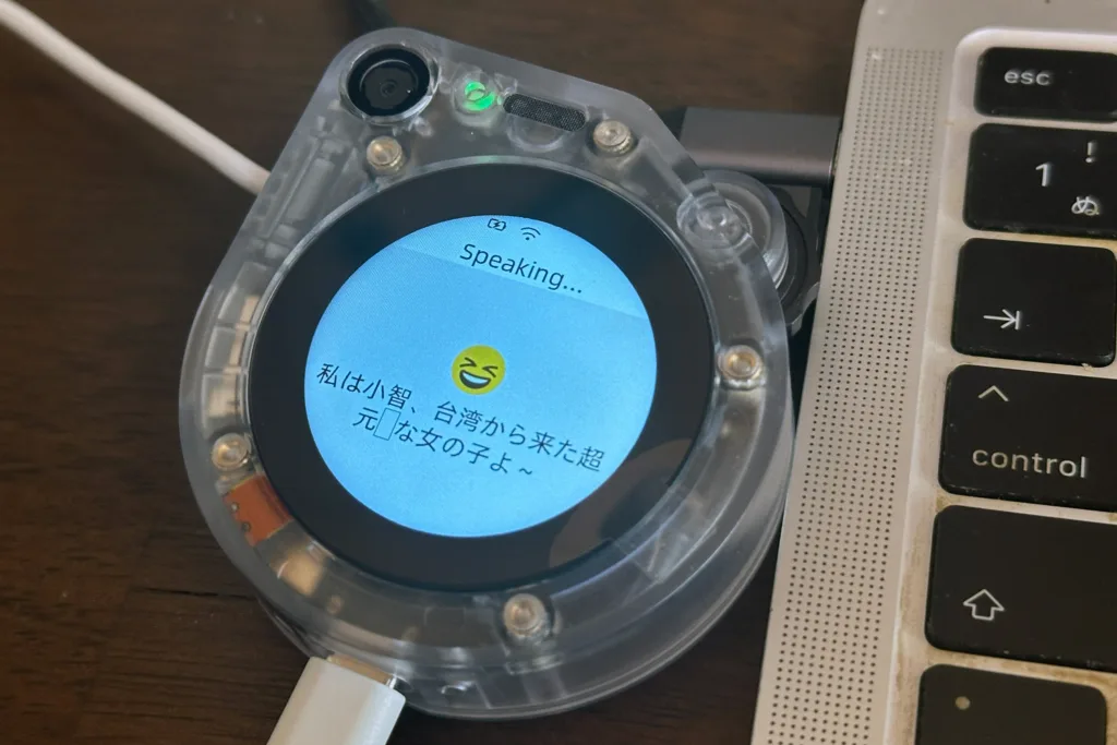 Circular transparent device on a desk displaying a blue screen that says 'Speaking...' with a laughing emoji, beside a laptop keyboard.