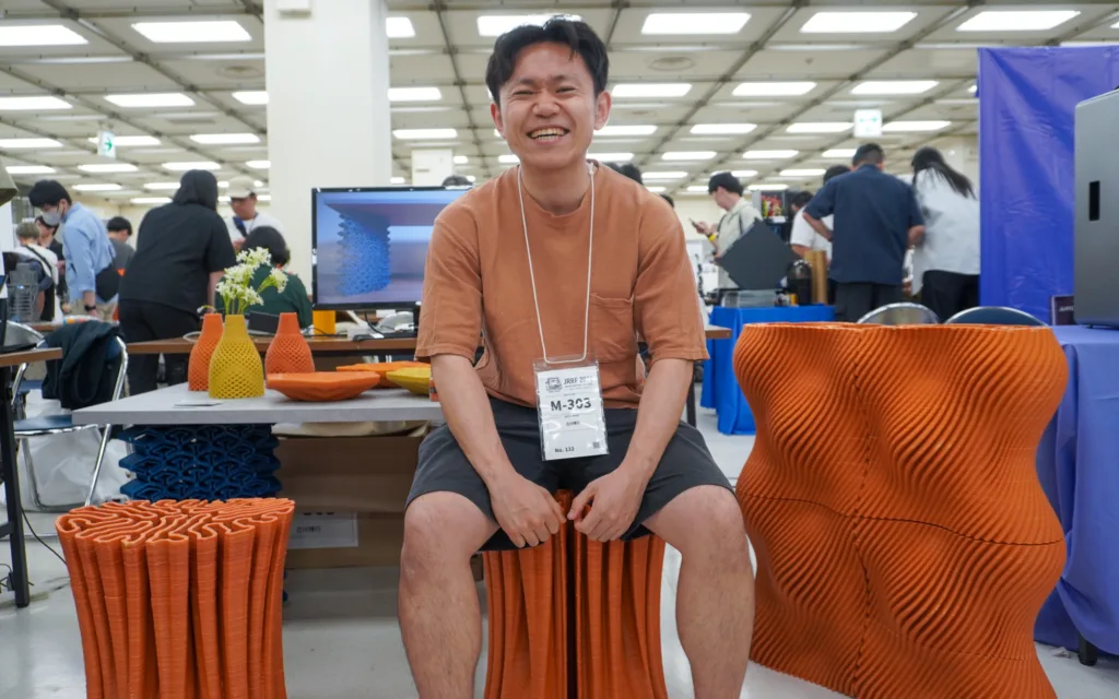 Smiling man wearing a badge sits on an orange corrugated stool at a technology expo, with displays and people in the background.