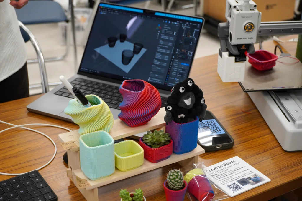 Laptop screen showing 3D modeling software on a wooden desk, with colorful knitted cable organizers and small succulents; a white 3D printer sits to the right.