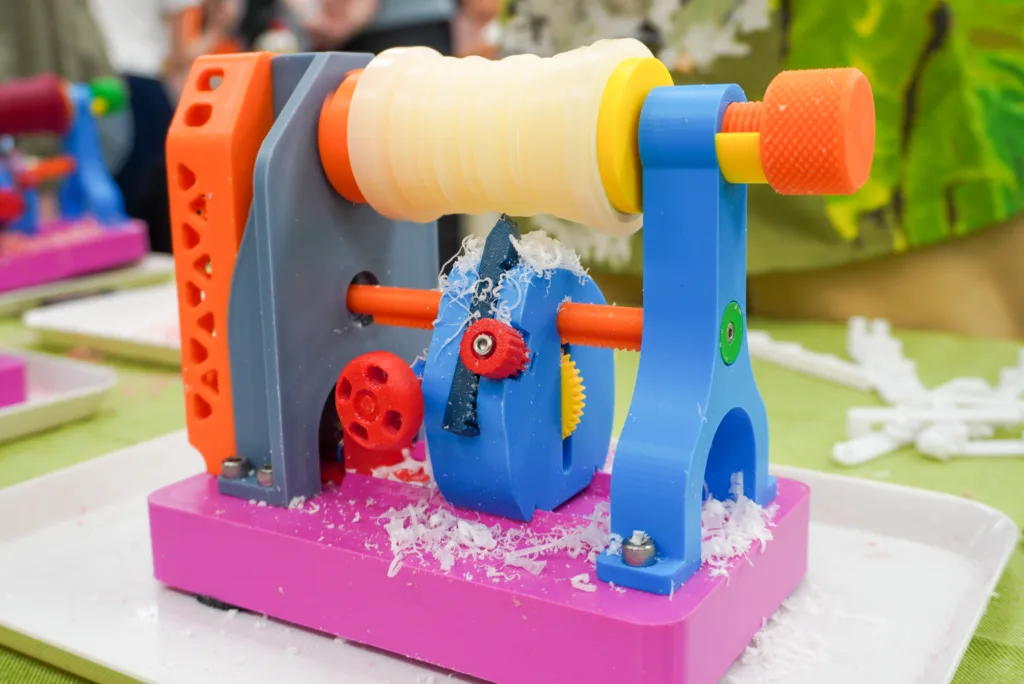 Close-up of a colorful plastic gear toy with a yellow roller, blue supports, orange crank knob, and pink base on a table.