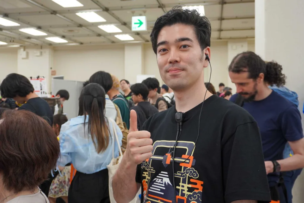 Man in a black graphic T-shirt giving a thumbs-up at a crowded indoor event, wearing a neckband headset.