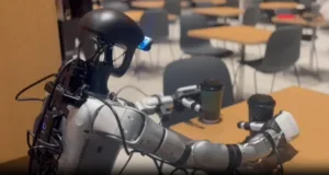 Close-up of a metallic robot with articulated arms on a classroom table, empty desks and chairs in the background.