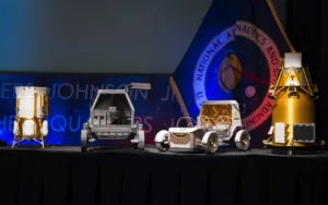 Models of space probes on a stage: white cylindrical lander, gray rover, white lunar buggy, and a gold lander, with a space-agency banner in the background.