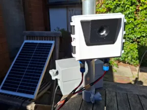 Outdoor solar-powered irrigation controller mounted on a pole, with red and black wires connected; a solar panel rests nearby on the ground.