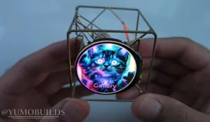 Close-up of a circular pendant showing a neon-color cat illustration and the word 'Gallery', held in a metal frame by fingers.