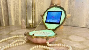 Open pink shell-shaped jewelry box with a digital screen showing a mermaid underwater; pearl necklaces spread across a marble surface.