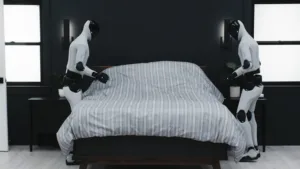 Two white humanoid robots stand on opposite sides of a bed, pulling up a striped gray duvet.