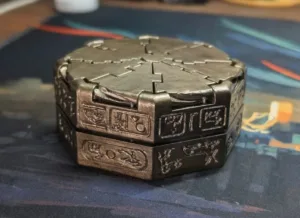 Metallic modular puzzle box with interlocking top panels and carved symbols on the sides, resting on a dark surface.
