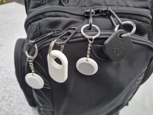 Close-up of a black backpack with multiple zippers, metal rings, and white circular tags attached to the front.