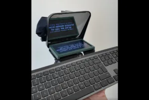 Compact fold-out display attached to a laptop, showing blue text on a dark screen in a green frame.