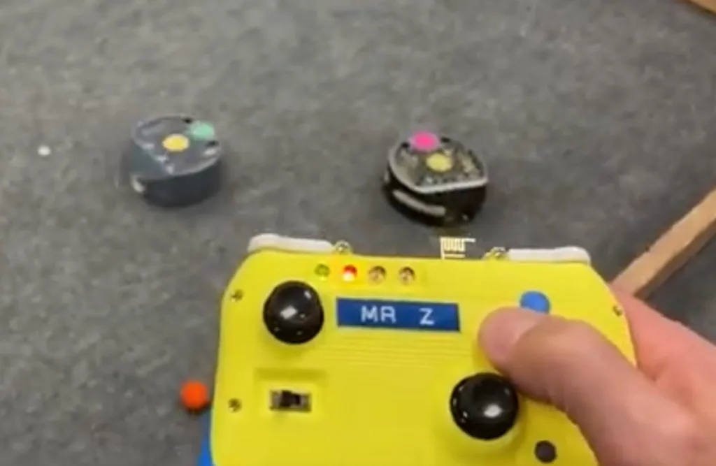 Yellow handheld remote labeled MR Z, with two joysticks, controlling two small round robots on a gray floor.