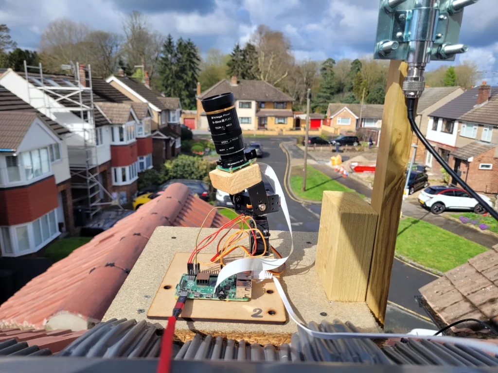 Camera lens and electronics rig installed on a wooden platform on a roof, with a suburban street in the background.